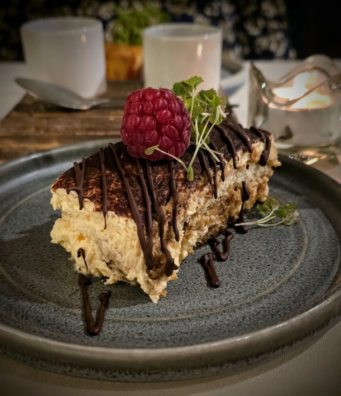 Layered tiramisu-style dessert with chocolate drizzle and a fresh raspberry, set against a candlelit table