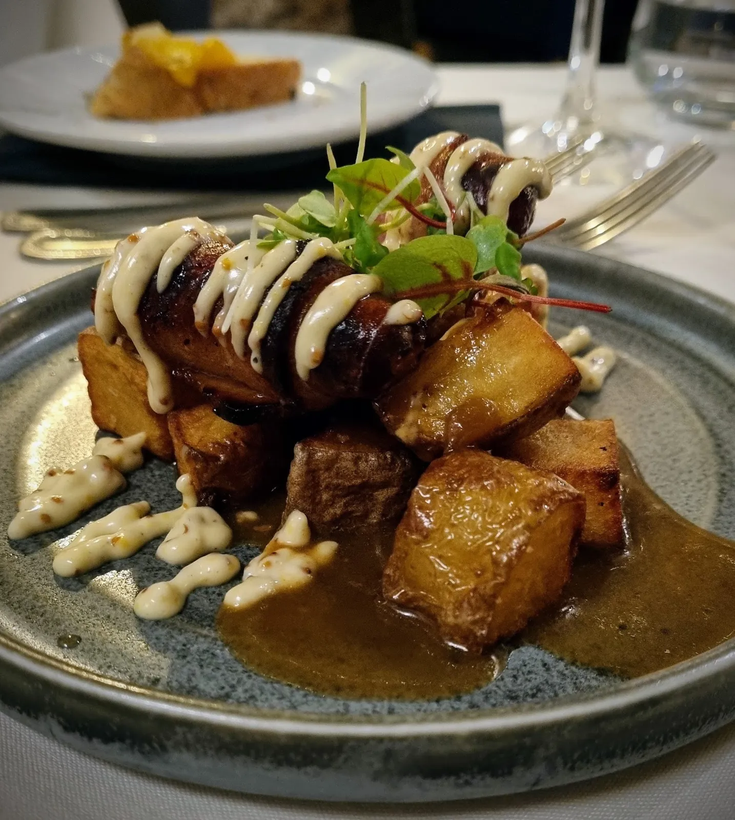 Bacon-wrapped pork with roasted potato cubes and a cream drizzle, plated under warm indoor light
