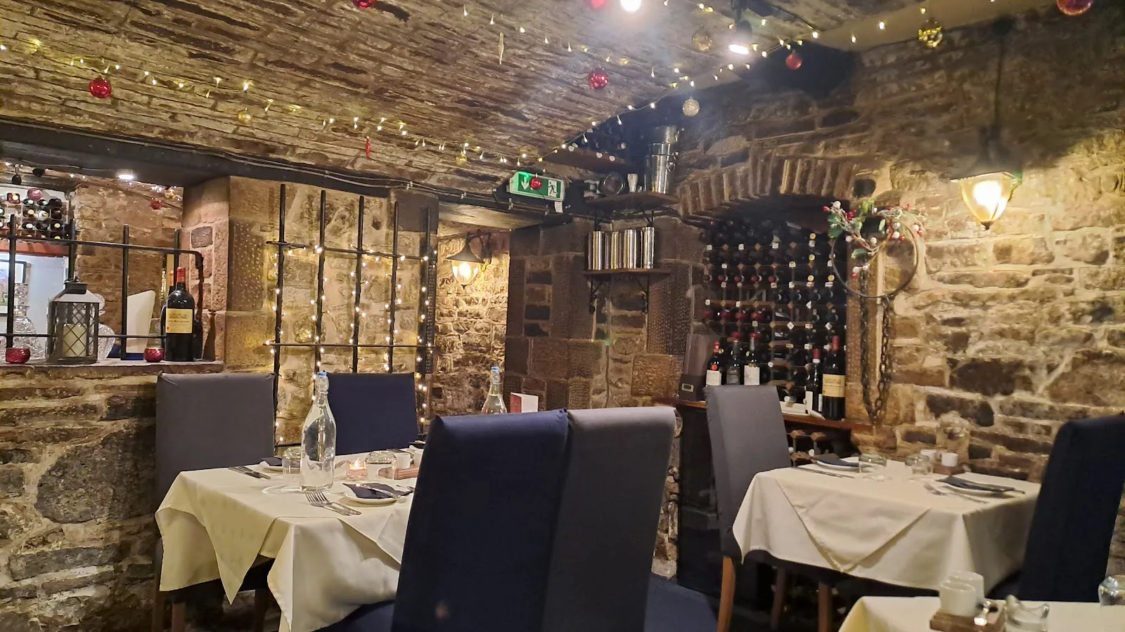 Stone-walled interior of Le Caveau's vaulted cellar, with white-linen tables, navy chairs, a wine rack, and fairy lights glowing beneath the ceiling