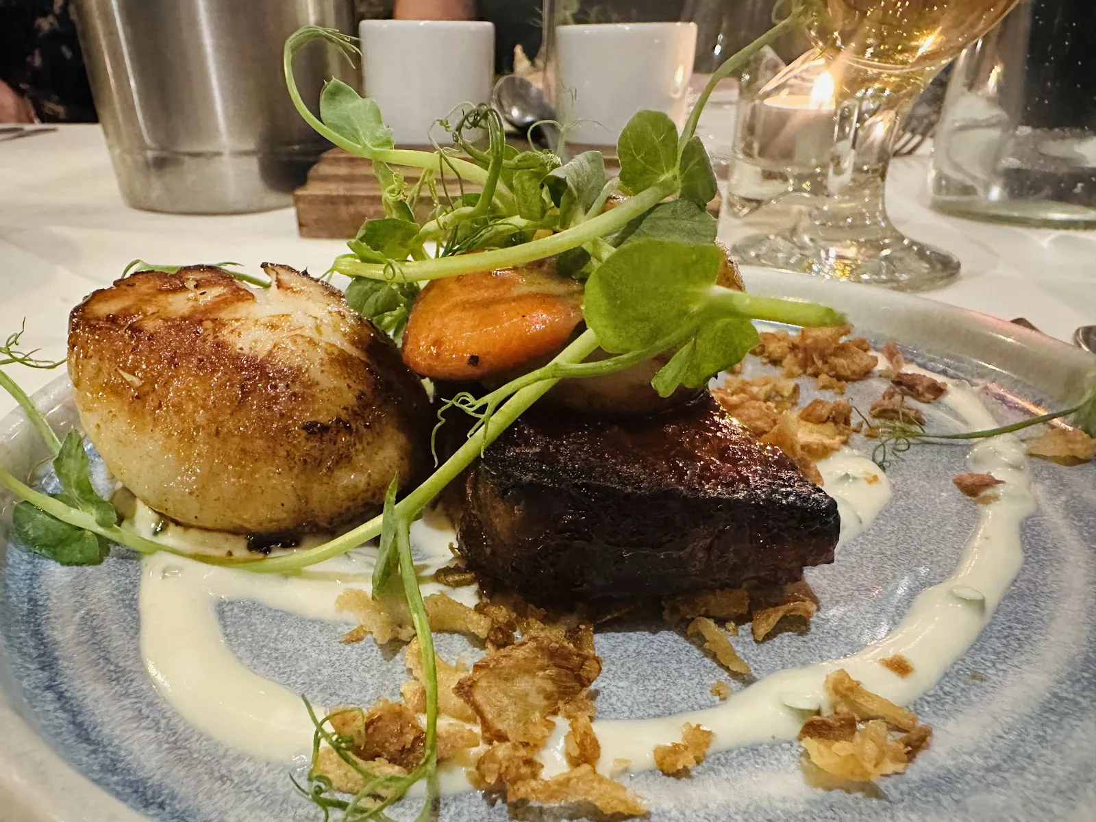 Seared scallop and slow-roasted pork belly plated with pea shoots and crispy shallots, lit by candlelight at the table